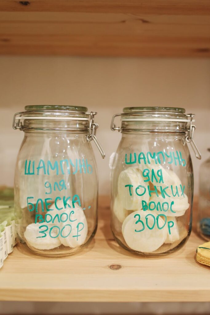 Zero waste shampoo bars in glass jars with Cyrillic labels on wooden shelf.